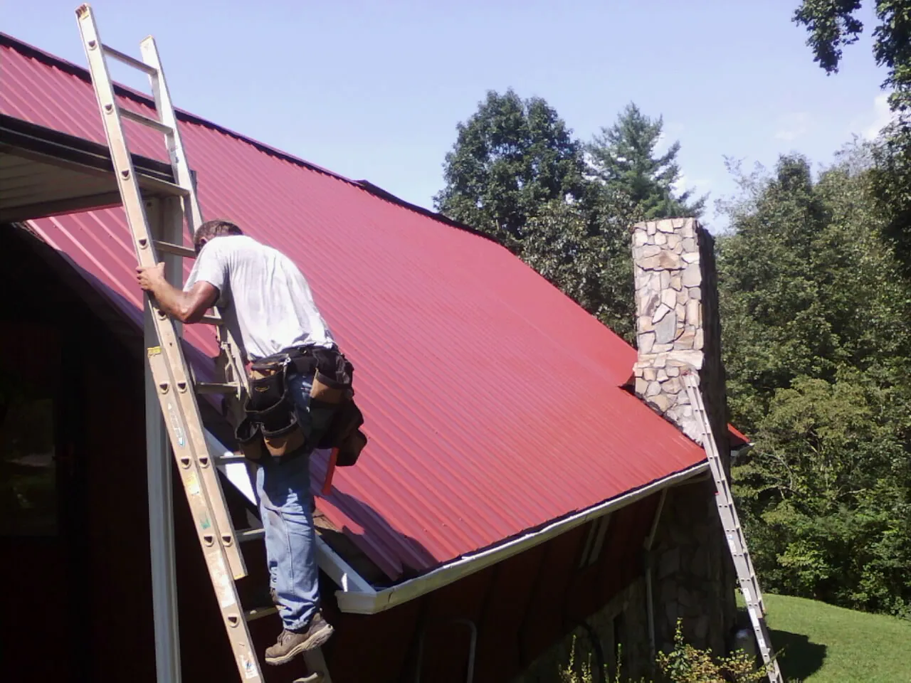 Roofer on a ladder beside a red metal roof and stone chimney — Soffit & Fascia Repair by Roofing Brothers in Blue Bell